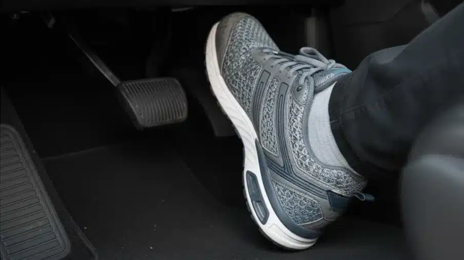 A person's foot in a grey and blue athletic shoe presses the accelerator pedal in a car, with the brake pedal visible to the left.