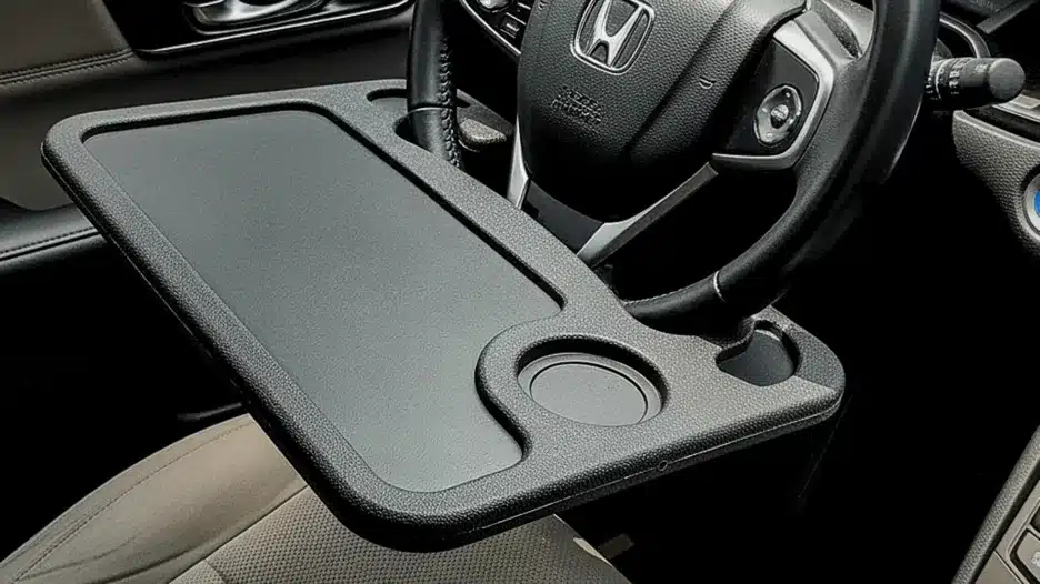 A close-up view of a black, plastic tray accessory with a flat surface and a cup holder, attached to the steering wheel of a car (appears to be a Honda) for use as a desk or eating surface when parked.