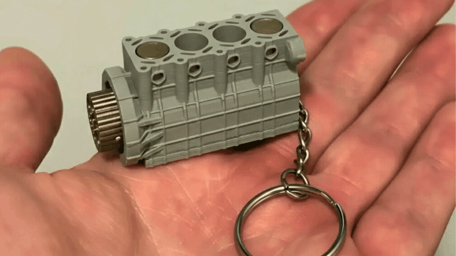 Close-up of a realistic 4-cylinder engine keychain resting on a hand, featuring visible pistons and a compact mechanical design for car enthusiasts.