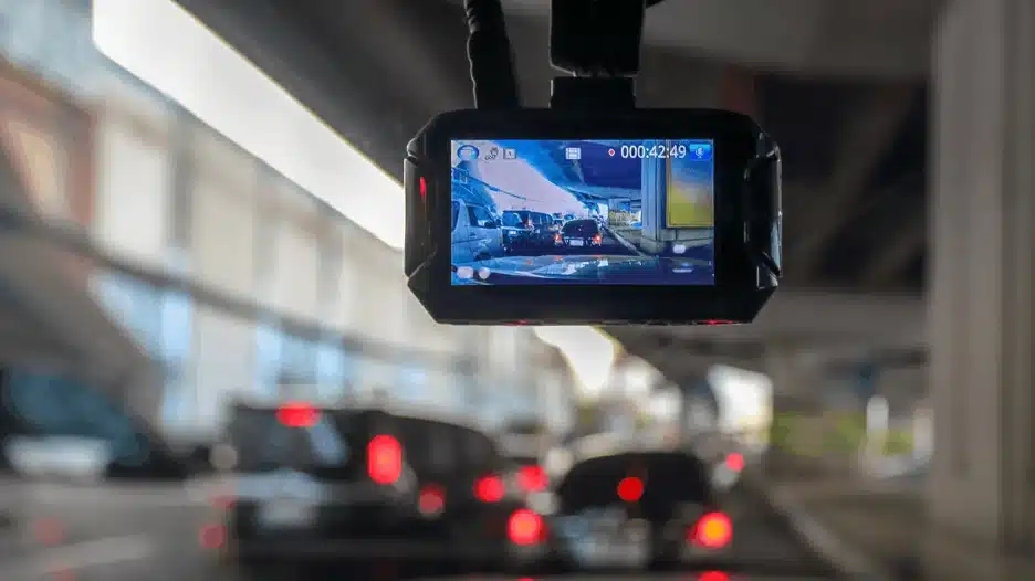 Dashboard camera mounted inside a car recording traffic on the road, showing live video footage on screen with vehicles and city roadway in view.