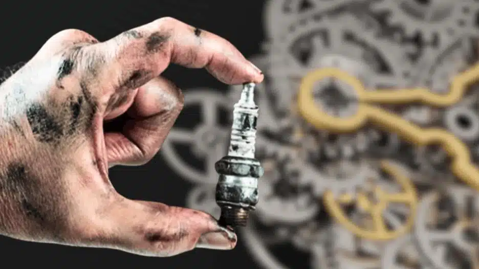 Hand holding a used spark plug with engine components blurred in background representing internal combustion engine ignition system.