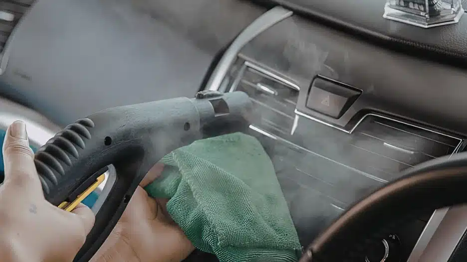 Person using a handheld steam cleaner and microfiber cloth to deep clean and sanitize a car dashboard and interior surfaces, removing dirt and buildup from vents and panels.