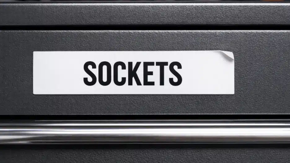 Close-up of a dark gray textured toolbox drawer with a clean white label strip reading “SOCKETS,” with the top-right corner of the label slightly lifted.